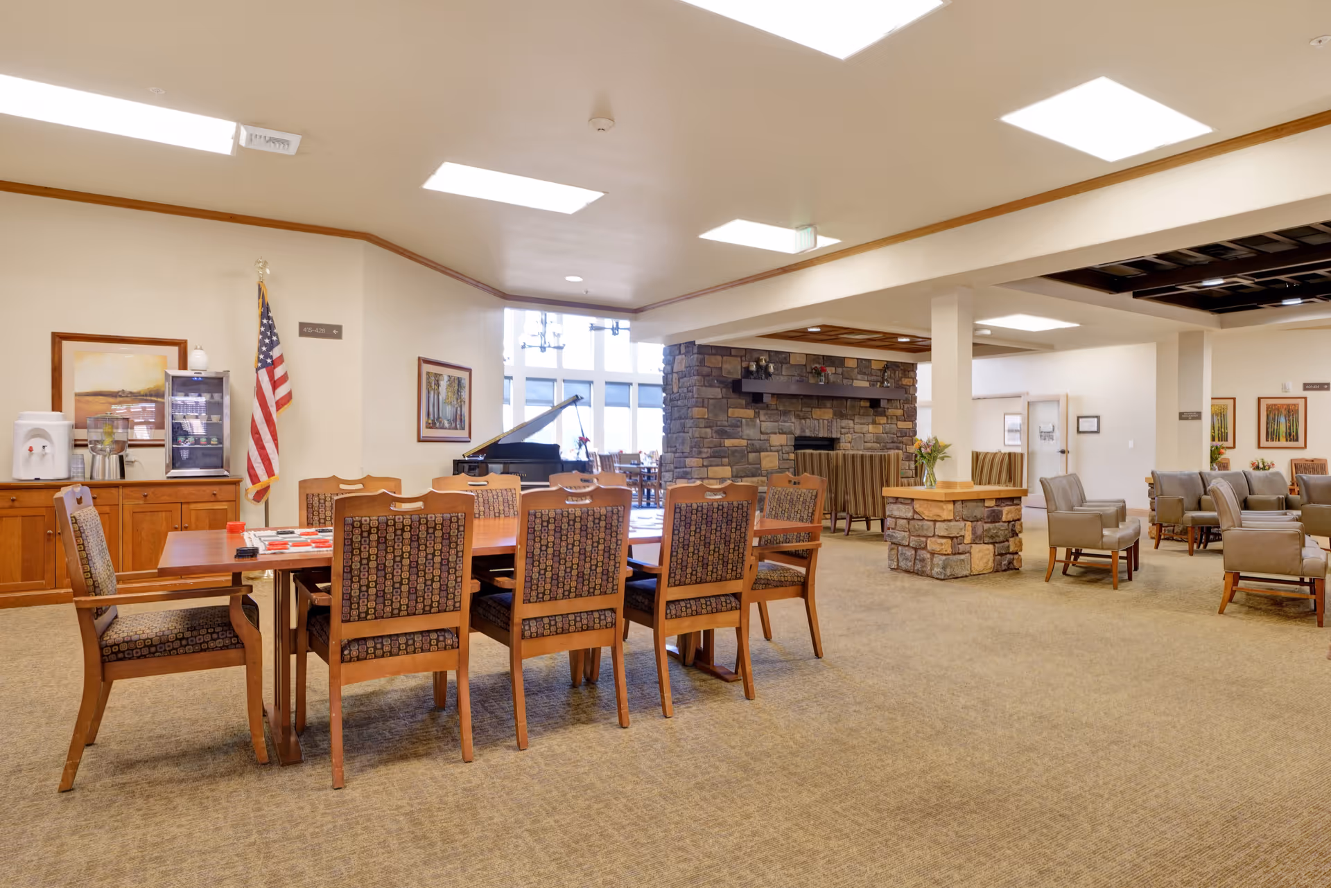 A spacious common area in a senior living facility featuring a large wooden table with eight patterned chairs, a stone fireplace, several armchairs arranged in small groups, a piano near large windows, and an American flag in the corner. The room is well-lit with ceiling lights and decorated with framed artwork and flowers on tables.