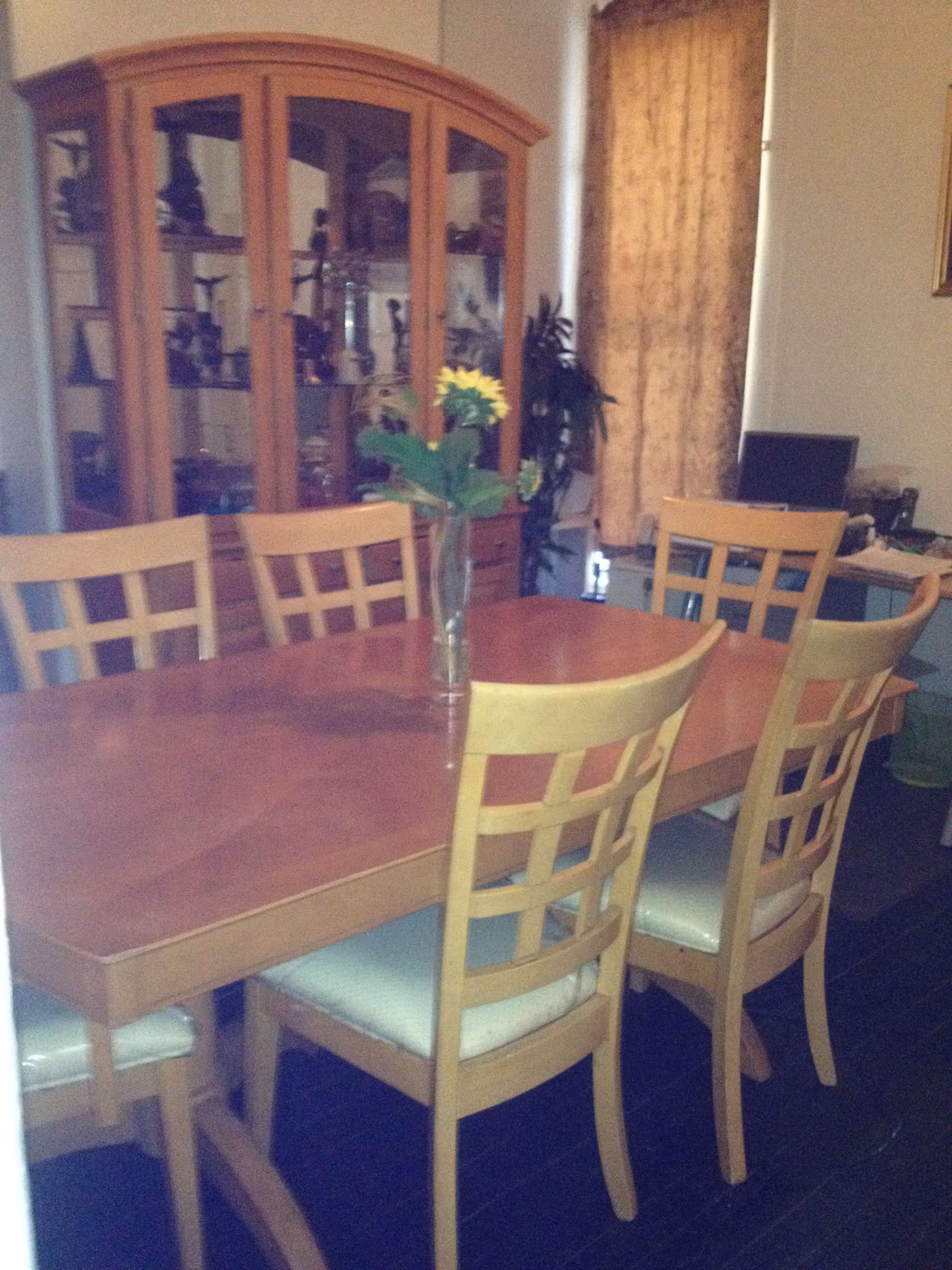 A dining room with a wooden dining table surrounded by six wooden chairs with cushioned seats. A glass vase with a yellow flower is placed on the table. In the background, there is a wooden china cabinet with glass doors displaying various items, a window with a curtain, and a desk with a laptop and other items.