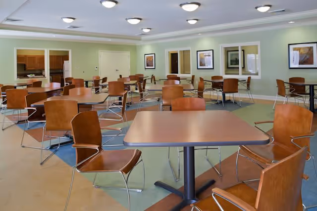 A spacious dining room with multiple square wooden tables and brown chairs arranged neatly. The room has light green walls, framed artwork, and ceiling lights. There is a kitchen area visible in the background with wooden cabinets and a refrigerator.