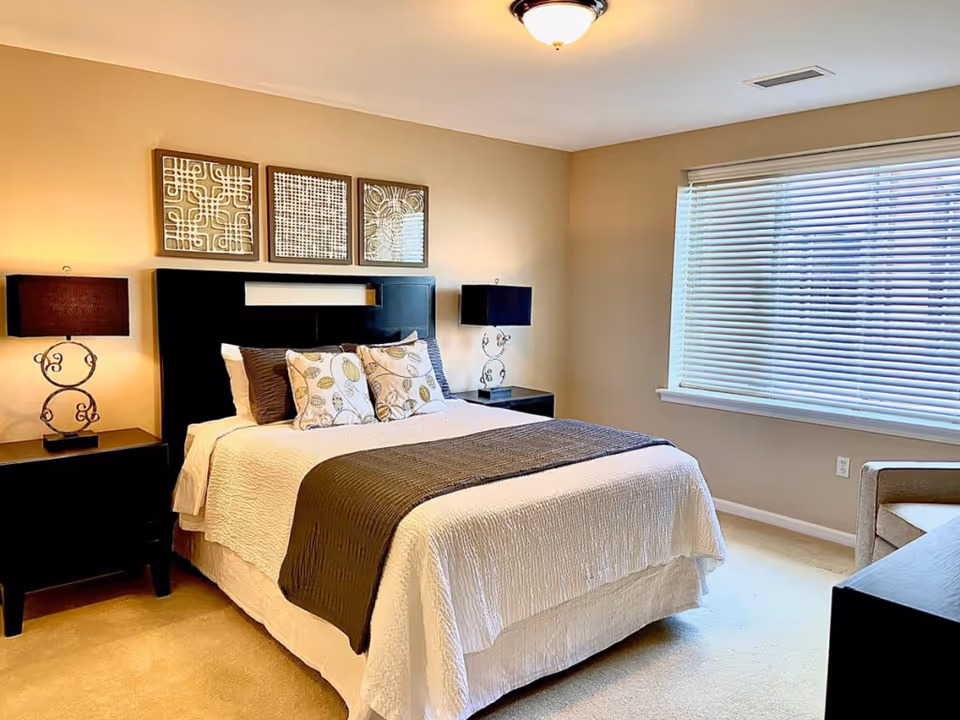 A neatly made bed with white and brown bedding in a bedroom. The bed has a black headboard and is flanked by two black nightstands, each with a lamp featuring a decorative base and dark lampshade. Above the bed are three framed abstract art pieces. There is a large window with white blinds on the right side of the room, and a beige armchair partially visible near the window. The walls are painted a light beige color.