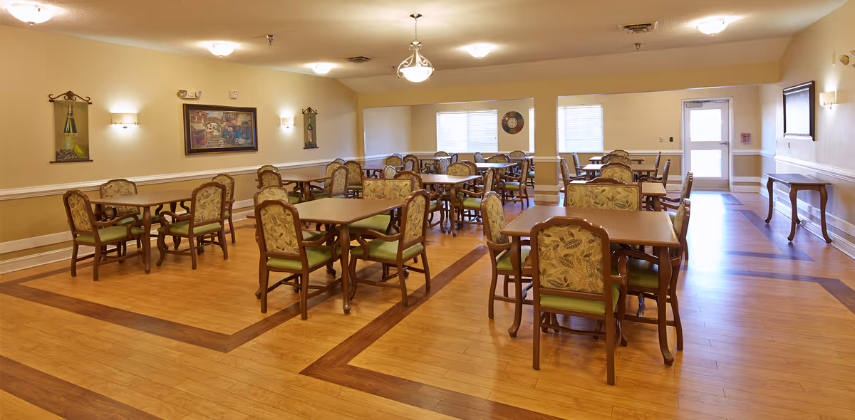 Spacious dining room with multiple wooden tables and upholstered chairs on patterned hardwood flooring, neutral walls with artwork and windows.