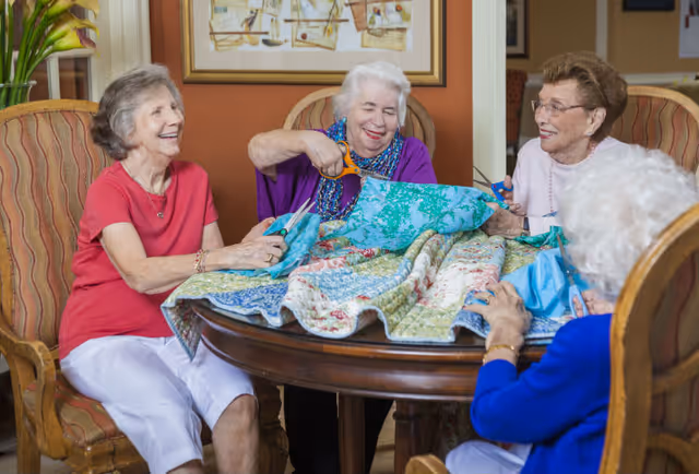 Four elderly women sitting around a wooden table, smiling and cutting pieces of colorful fabric, engaging in a group craft activity in a cozy indoor setting with framed artwork and plants in the background.