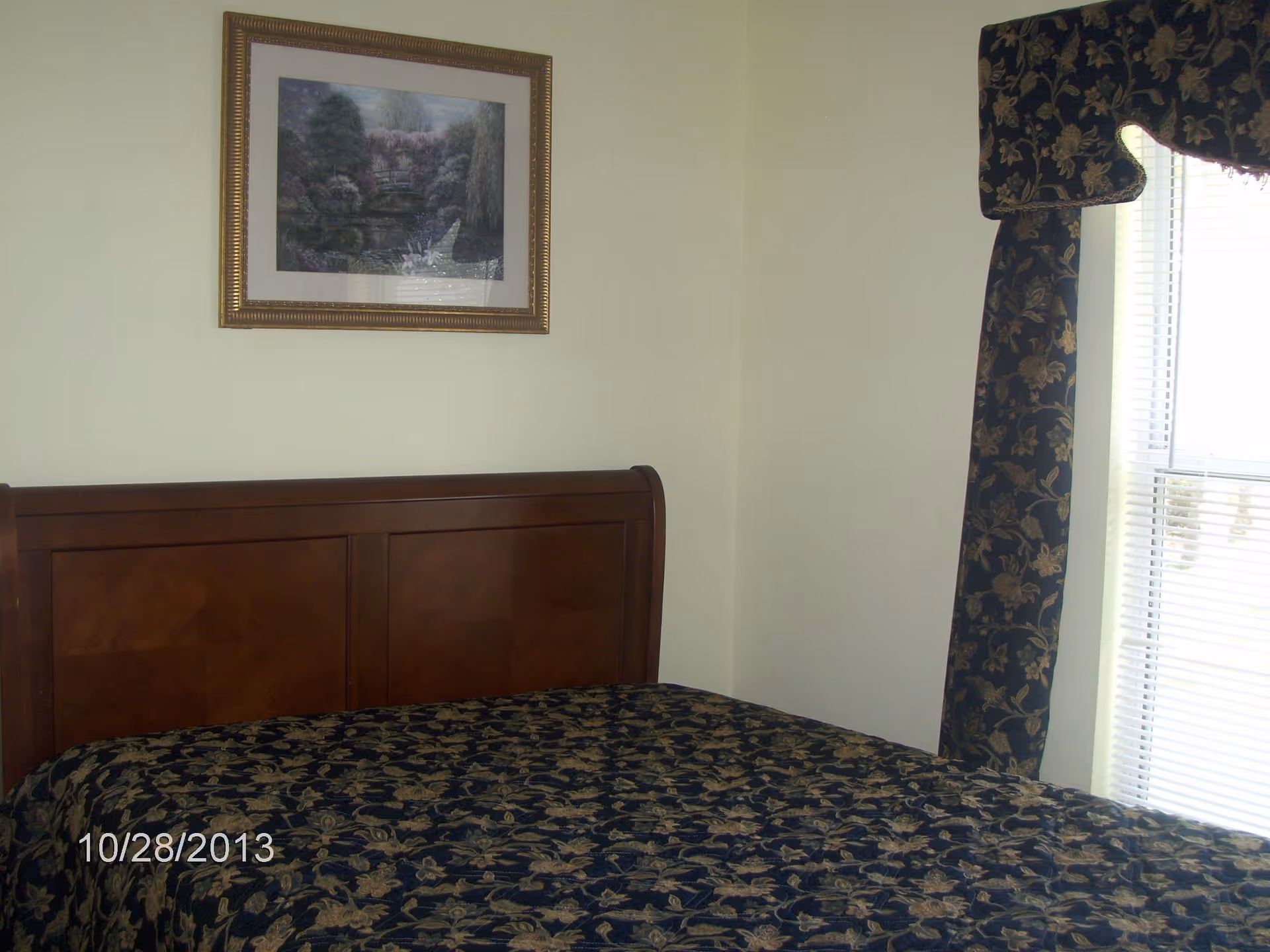 A bedroom with a wooden headboard bed covered with a dark floral patterned bedspread. There is a framed landscape painting on the wall above the bed and a window with floral curtains to the right.