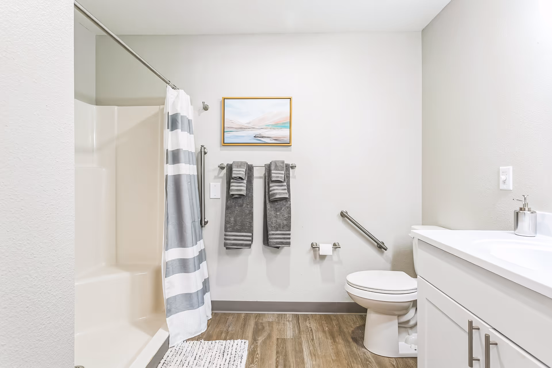 A clean and modern bathroom featuring a white shower with a gray and white striped curtain, two gray towels hanging on a towel rack, a toilet with a grab bar beside it, a white vanity with a sink and soap dispenser, light wood flooring, and a framed abstract painting on the wall.