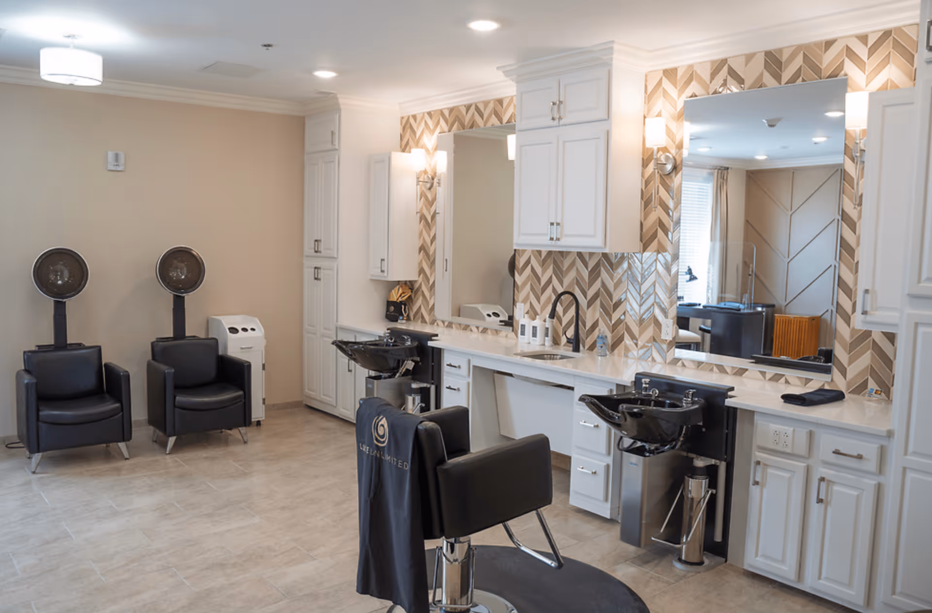 Interior view of a modern hair salon area with two black salon chairs facing mirrors, two black hair drying chairs with hooded dryers, white cabinetry, and a chevron-patterned tile backsplash.
