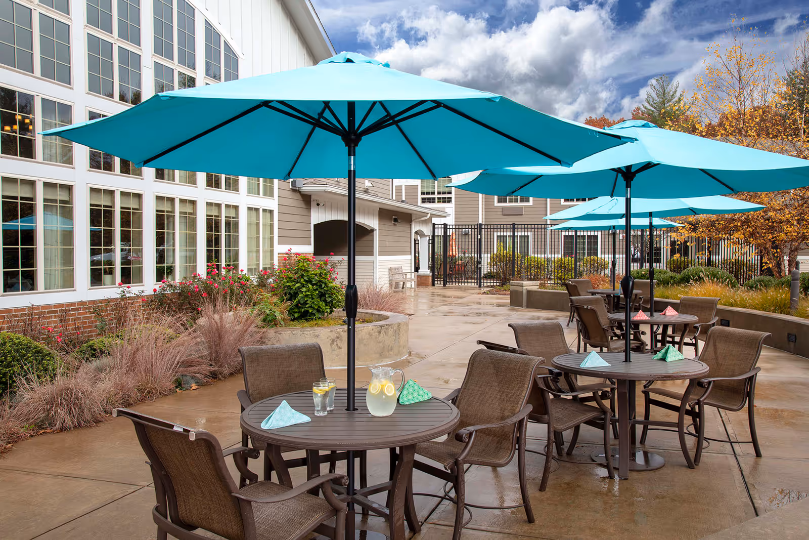 Outdoor patio area at Farmington Station Senior Living with multiple round tables, each shaded by large turquoise umbrellas. Each table has four brown chairs and is set with folded napkins and pitchers of lemonade. The patio is surrounded by landscaping with bushes and ornamental grasses, and the building with large windows is visible in the background under a partly cloudy sky.
