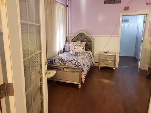 A bedroom with a single bed featuring a tufted headboard and patterned bedding. There is a small nightstand with a drawer next to the bed. The walls are painted light purple on the upper half and white on the lower half. The floor is wooden, and there is a door with glass panels partially open leading into the room. Another doorway leads to a hallway or adjacent room.