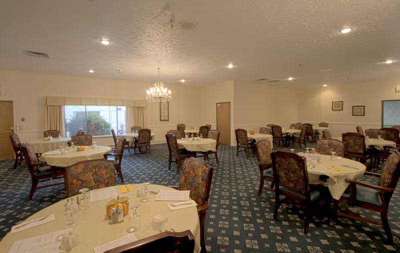 A dining room with multiple round tables covered with white tablecloths, each surrounded by floral upholstered chairs. The tables are set with menus, glasses, napkins, and small decorative pumpkins. A chandelier hangs from the ceiling, and there is a large window with beige curtains letting in natural light. The carpet is patterned in blue and white.