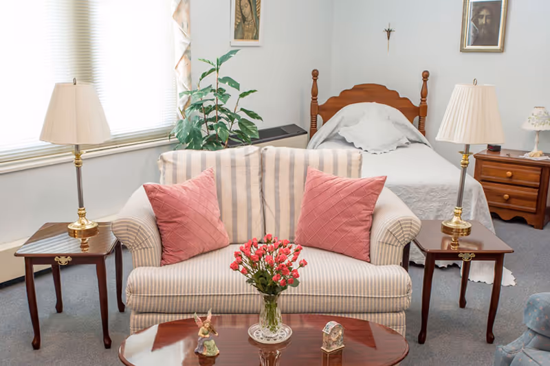 A cozy senior facility room with a striped loveseat and coffee table in front of a single bed and nightstands.