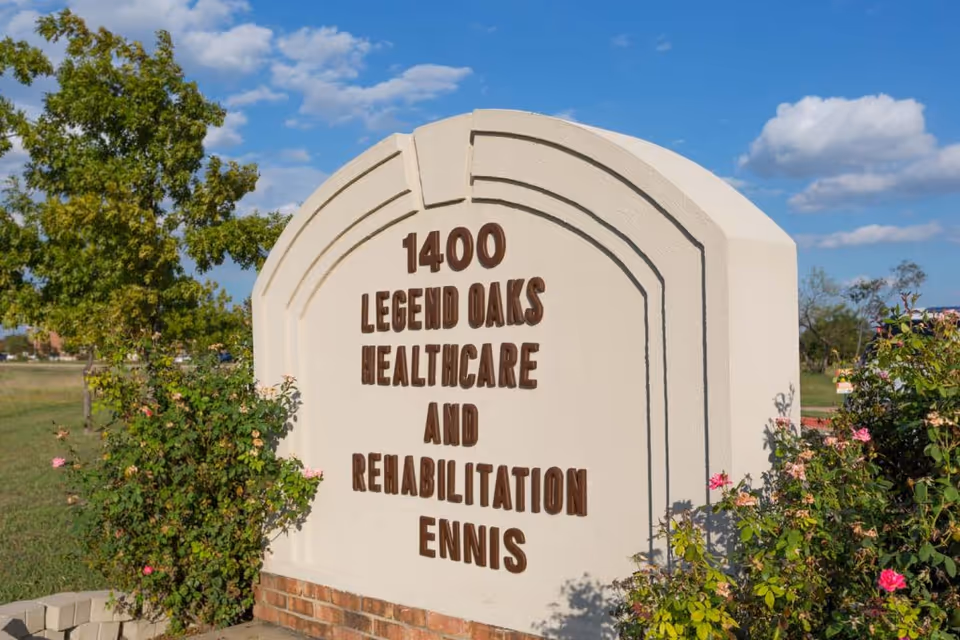 A large beige stone sign with brown letters that reads '1400 Legend Oaks Healthcare and Rehabilitation Ennis' surrounded by green bushes and pink flowers under a blue sky with scattered clouds.