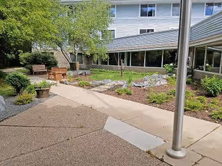 Outdoor courtyard area at a senior living facility with a paved walkway, garden beds with plants and rocks, benches, and a building with large windows in the background.