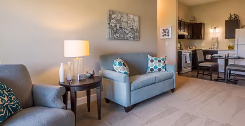 Open-plan living area featuring a loveseat and armchair with patterned pillows, a side table and lamp, and a view into a small kitchen and dining area.