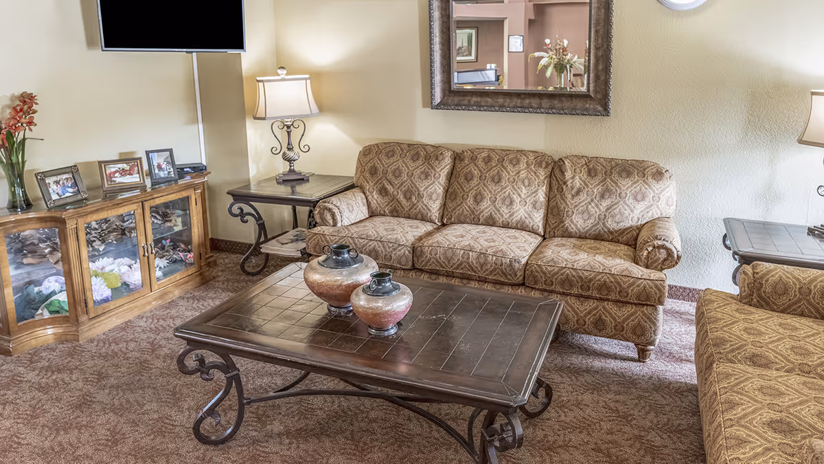 A cozy living room with patterned upholstered sofas, a dark wooden coffee table with two decorative vases, a side table with a lamp, a wooden cabinet with glass doors displaying flowers and framed photos, and a wall-mounted flat-screen TV. A large mirror hangs on the wall above the sofa.