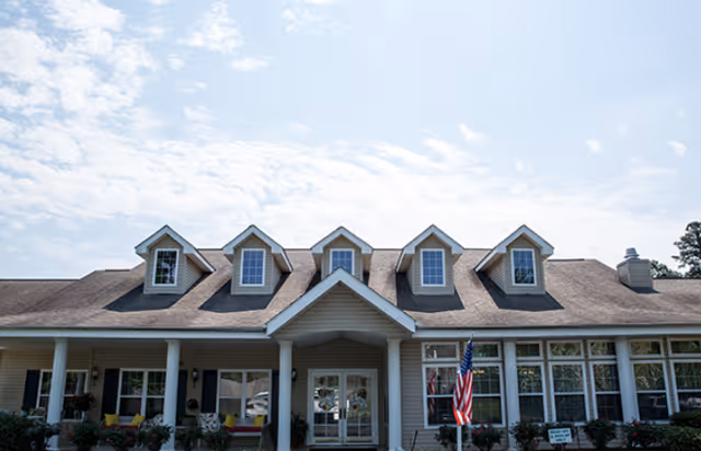 Front exterior view of a single-story building with a pitched roof and five dormer windows. The entrance has a covered porch supported by white columns, with an American flag displayed near the door. There are multiple windows along the front and some landscaping with bushes and flowers.