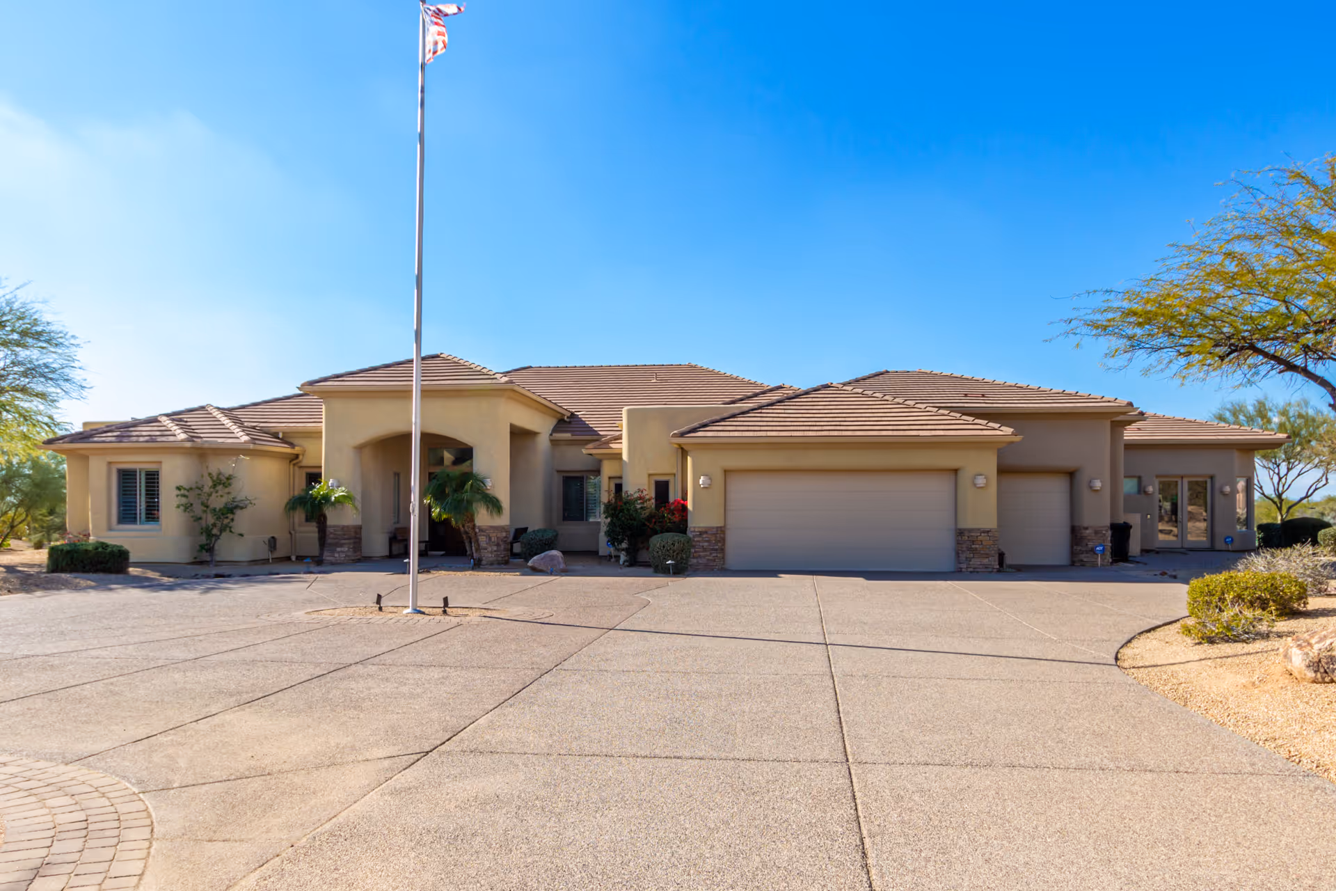 Front exterior view of a single-story building with a beige stucco finish and a tiled roof. There is a large driveway in front, a flagpole with an American flag, and desert landscaping with small trees and bushes under a clear blue sky.