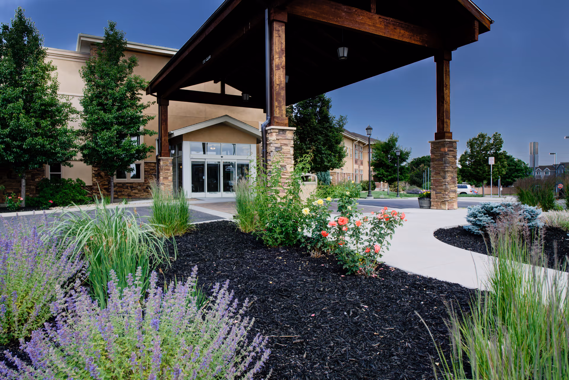 Entrance of a senior living facility named Gardens on Quail with a covered driveway supported by stone pillars, surrounded by landscaped garden beds with various plants and flowers, and a clear blue sky.