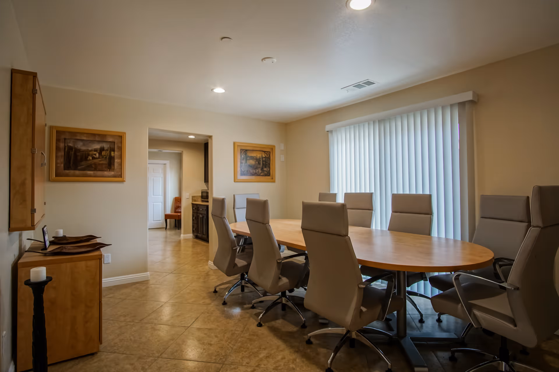 A well-lit dining room with a large oval wooden table surrounded by eight beige cushioned chairs on wheels. The room has beige tiled flooring and cream-colored walls adorned with two framed paintings. A large window with vertical blinds allows natural light to enter. There is a wooden cabinet on the left side with decorative items on top and a candle holder on the floor.