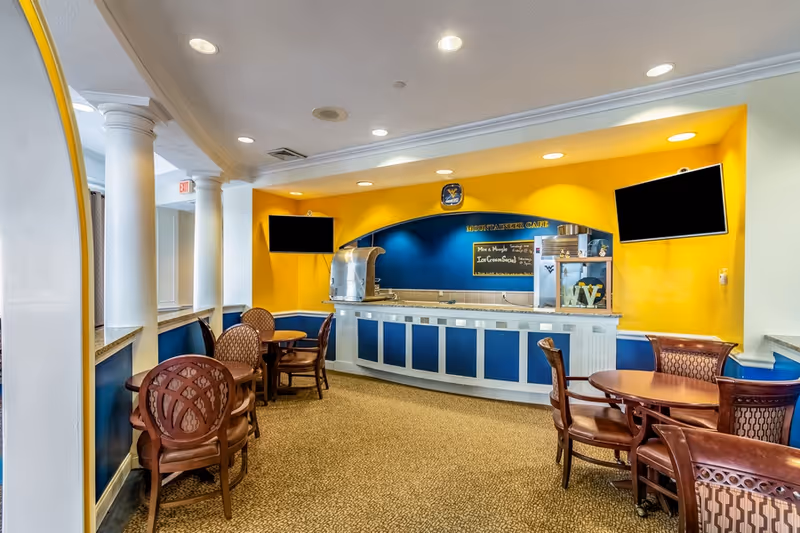 Bright indoor café/dining area with a blue-and-yellow service counter and several round tables and chairs.