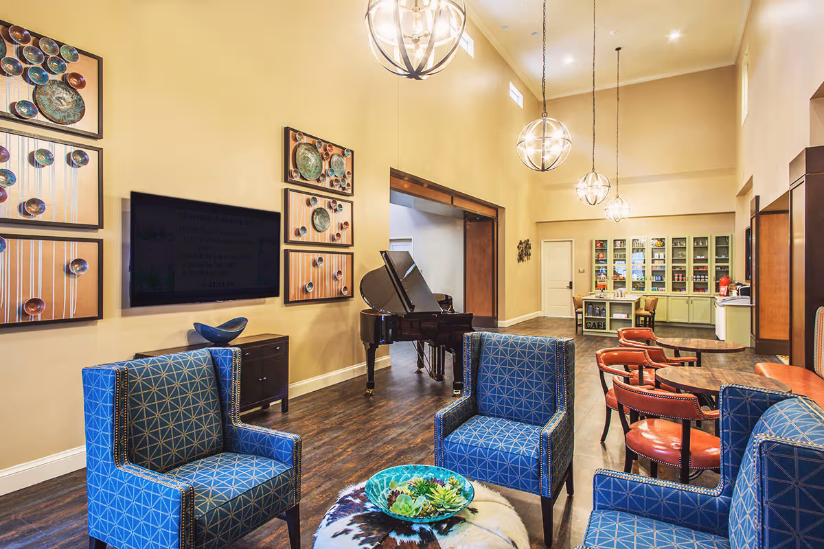 A spacious and well-lit common area with high ceilings featuring modern spherical pendant lights. The room includes blue patterned armchairs, a round table with red chairs, a black grand piano, and a wall-mounted flat screen TV. Decorative wall art is displayed above the TV, and a cabinet with glass doors filled with various items is visible in the background.