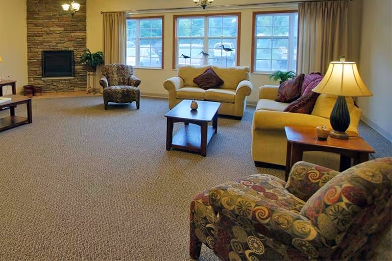 Bright common living room with sofas, patterned armchairs, a coffee table, lamp, and a stone fireplace beneath large windows.