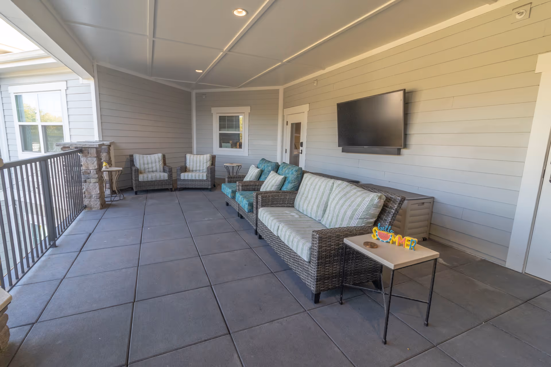 Covered outdoor seating area with wicker sofas and chairs, side tables, and a wall-mounted TV on a tiled balcony.