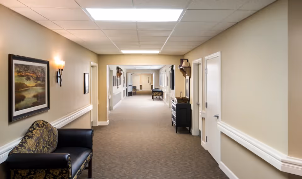 A well-lit hallway in a senior living facility with beige walls, carpeted floor, and white doors on the right side. There is a patterned armchair on the left, a framed landscape painting on the wall, and a small black cabinet with decorative items on the right. The hallway extends into the distance with additional seating and artwork visible.
