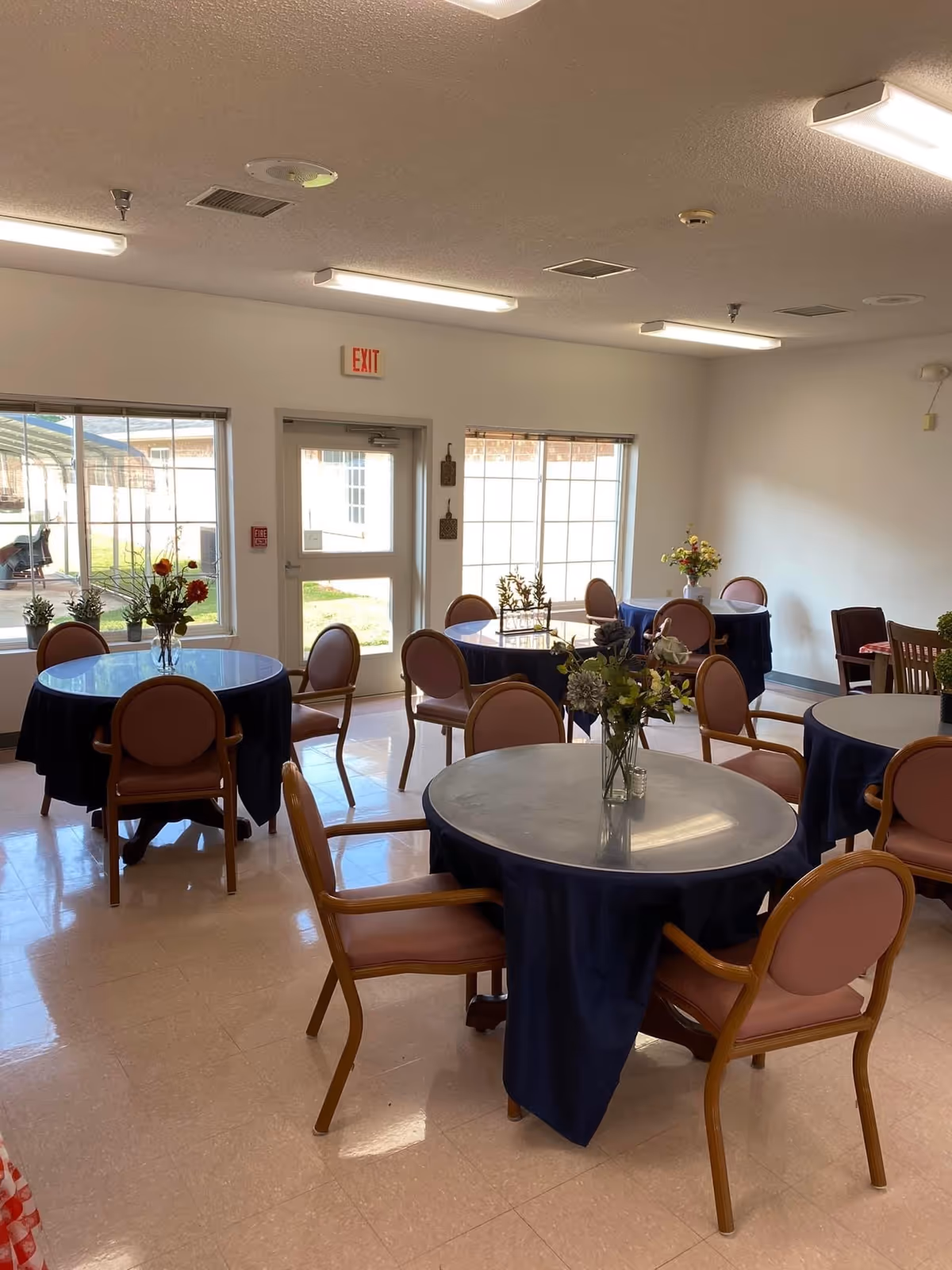 A dining room with several round tables covered with dark blue tablecloths and surrounded by wooden chairs with pink cushions. Each table has a vase with flowers. The room has large windows letting in natural light and an exit door with an exit sign above it.