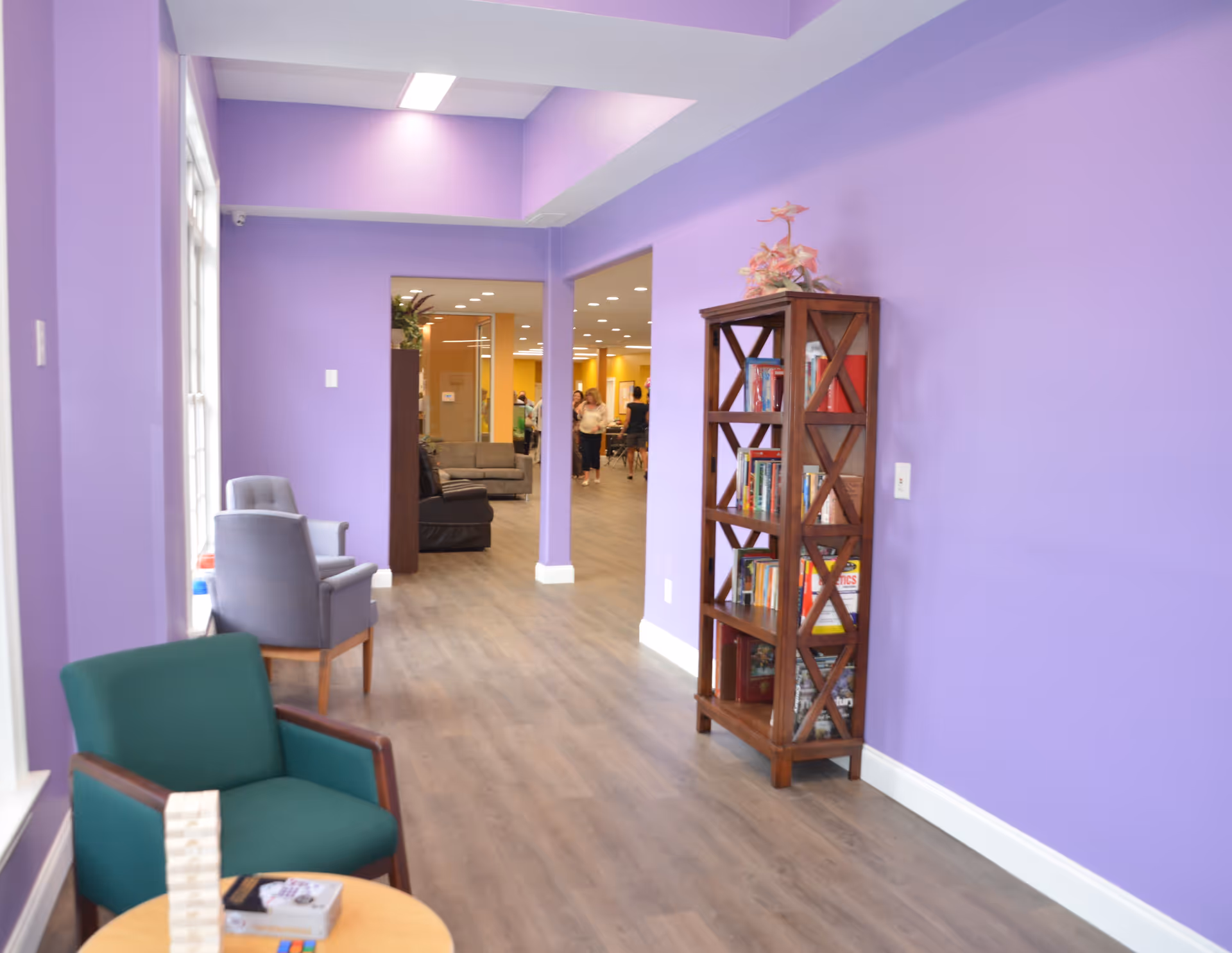 A bright purple-walled lounge corridor with chairs, a small table, and a wooden bookshelf opening into a seating area.