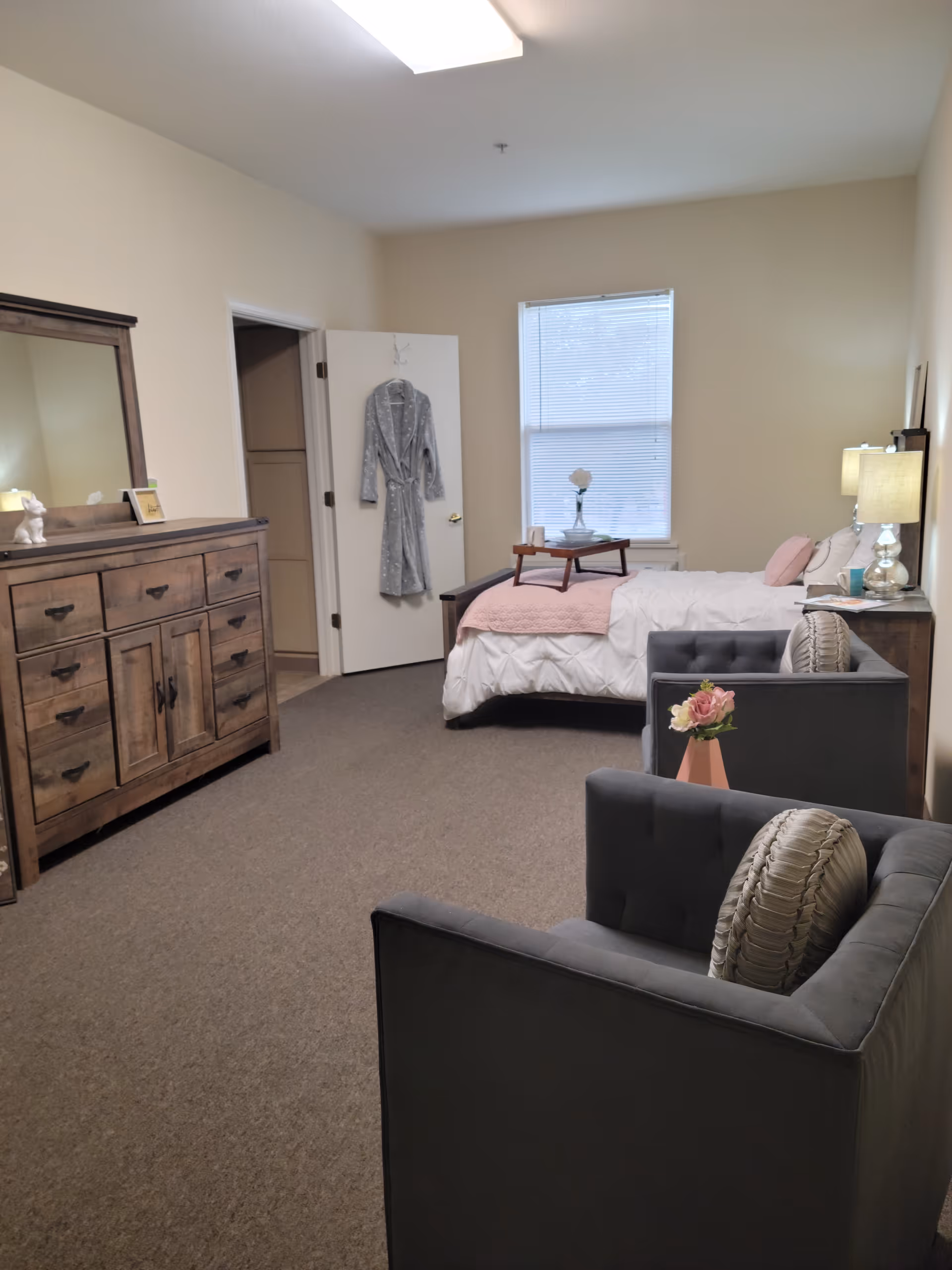 A cozy senior living bedroom with a bed covered in white and pink bedding, a wooden dresser with a mirror, two gray armchairs with decorative pillows, a small table with a flower vase, and a robe hanging on the door. The room has beige walls and carpeted floor.