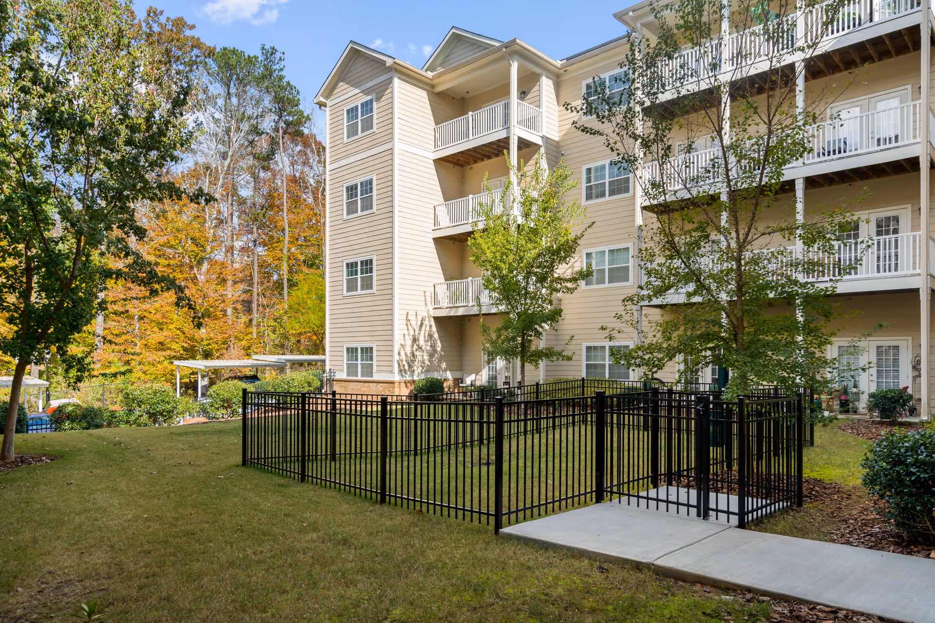 Exterior view of a multi-story senior living facility building with beige siding and white balconies. The building is surrounded by green grass, trees with autumn foliage, and a black metal fenced area with a concrete pathway leading to it. The sky is clear and blue.
