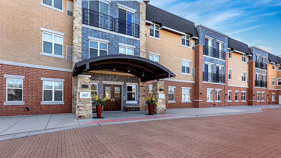 Exterior view of a multi-story senior living facility building with a covered entrance supported by stone pillars, large windows, and decorative balconies. The building features a mix of brick and stone facade with a paved driveway in front.