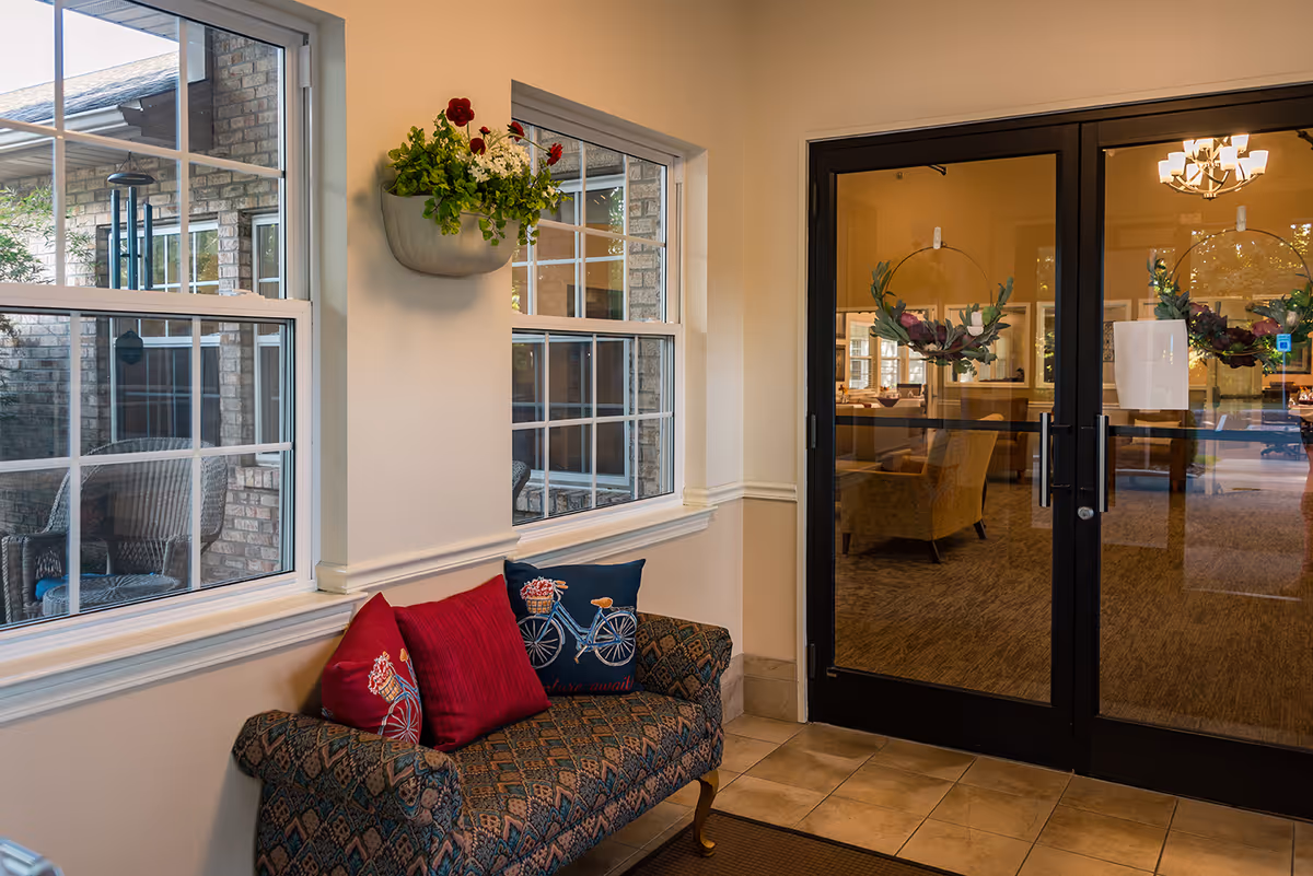 Cozy entryway with a patterned bench and decorative pillows beneath windows, a wall planter, and double glass doors leading into a common area.