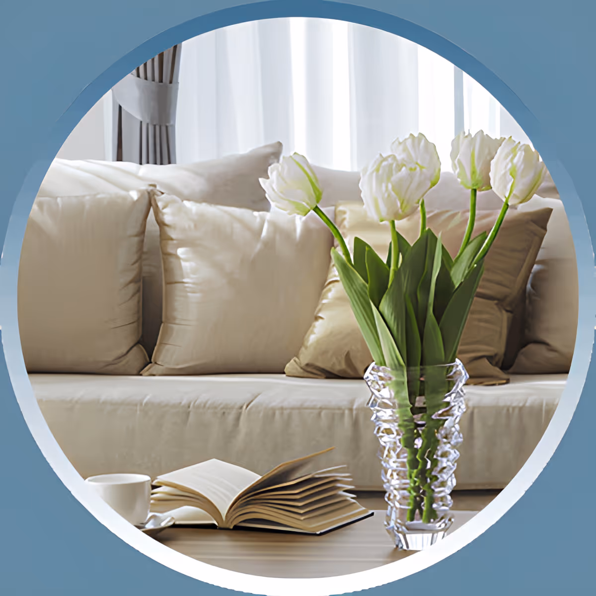 A cozy living room scene featuring a beige sofa with multiple cushions, a wooden coffee table with an open book and a white cup, and a clear glass vase holding white tulips in the foreground.