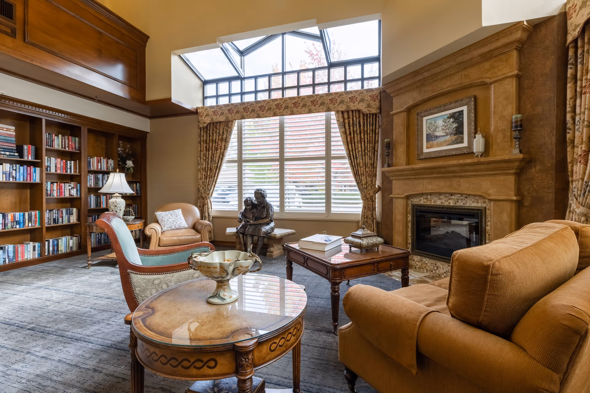 A cozy senior living facility common area with a large window featuring floral curtains, a stone fireplace with a framed painting above it, comfortable upholstered chairs and a sofa, a wooden coffee table with books and decorative items, a side table with a lamp, a bookshelf filled with books, and a bronze sculpture of two children sitting on a bench near the window.