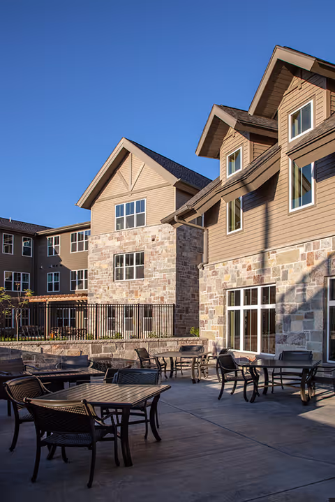 Outdoor patio area with multiple tables and chairs in front of a multi-story building with stone and beige siding under a clear blue sky.