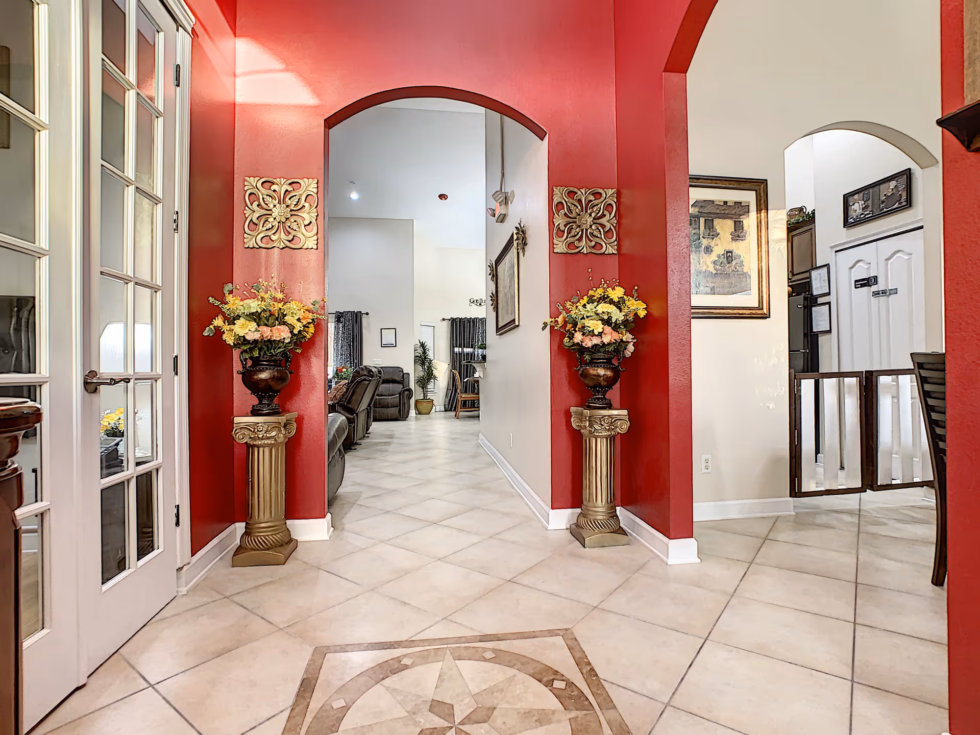 Entrance hallway with red arched walls, decorative pedestals holding flower arrangements, tiled floor and a view into a living room.