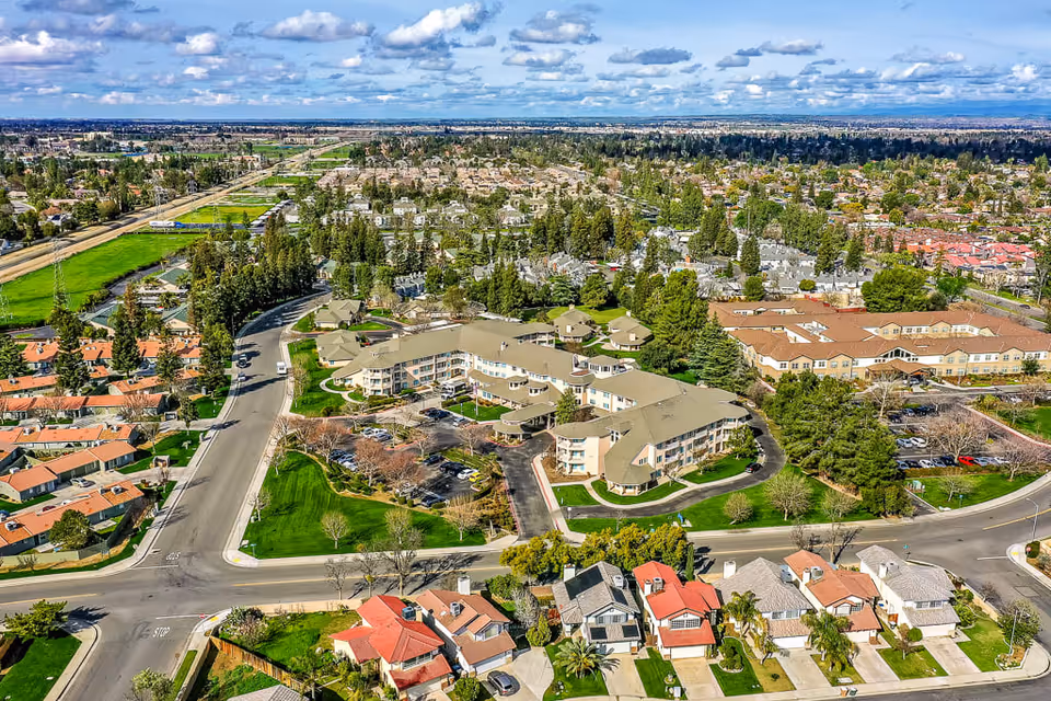 Aerial view of a suburban neighborhood featuring a large senior living facility with multiple connected buildings surrounded by green lawns, trees, and parking lots. Residential houses with red and gray roofs are visible in the foreground, and a wide road runs alongside the facility. The sky is partly cloudy with scattered clouds.