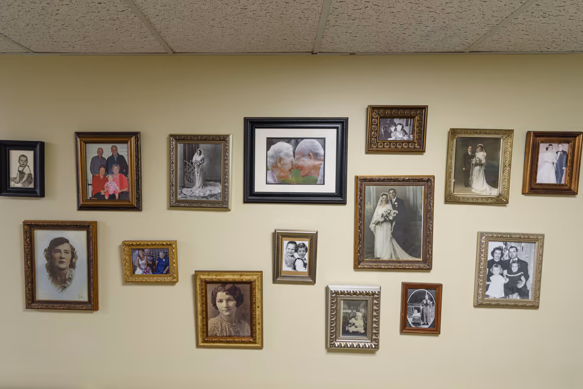 A beige wall decorated with multiple framed photographs, mostly vintage black and white portraits and wedding photos, along with a few color pictures of elderly people and families.