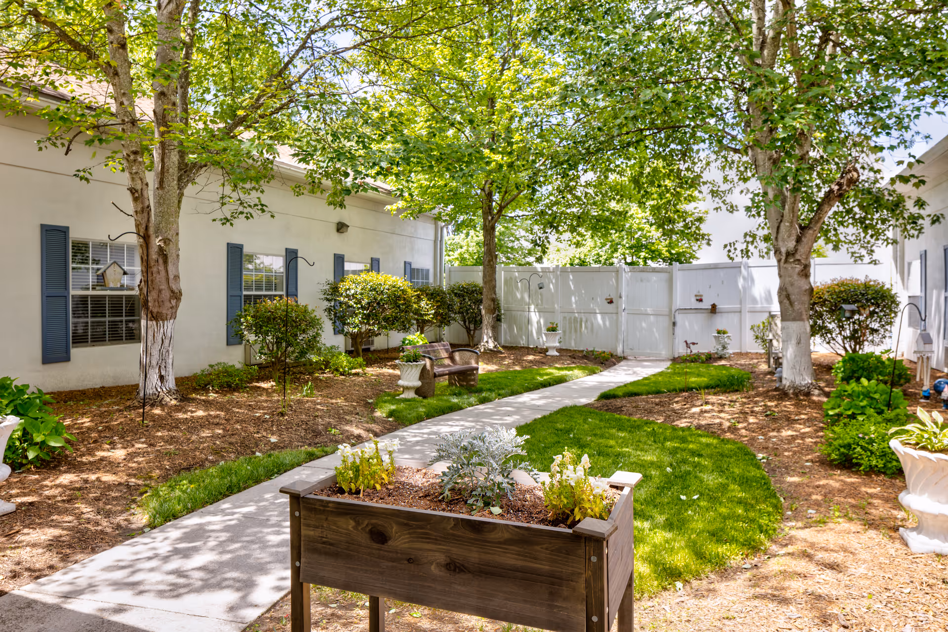 A peaceful outdoor garden area at TerraBella Little Avenue featuring a concrete pathway winding through green grass and mulched flower beds. There are trees providing shade, bushes along the building with blue shutters, a wooden planter box with plants in the foreground, and a bench for seating near the back white fence.