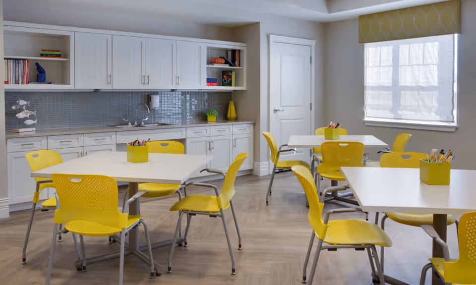 A bright and clean room with several white tables surrounded by yellow chairs. The room has a kitchenette area with white cabinets, a sink, and a tiled backsplash. There are small containers with art supplies on each table. A window with a yellow patterned valance lets in natural light.