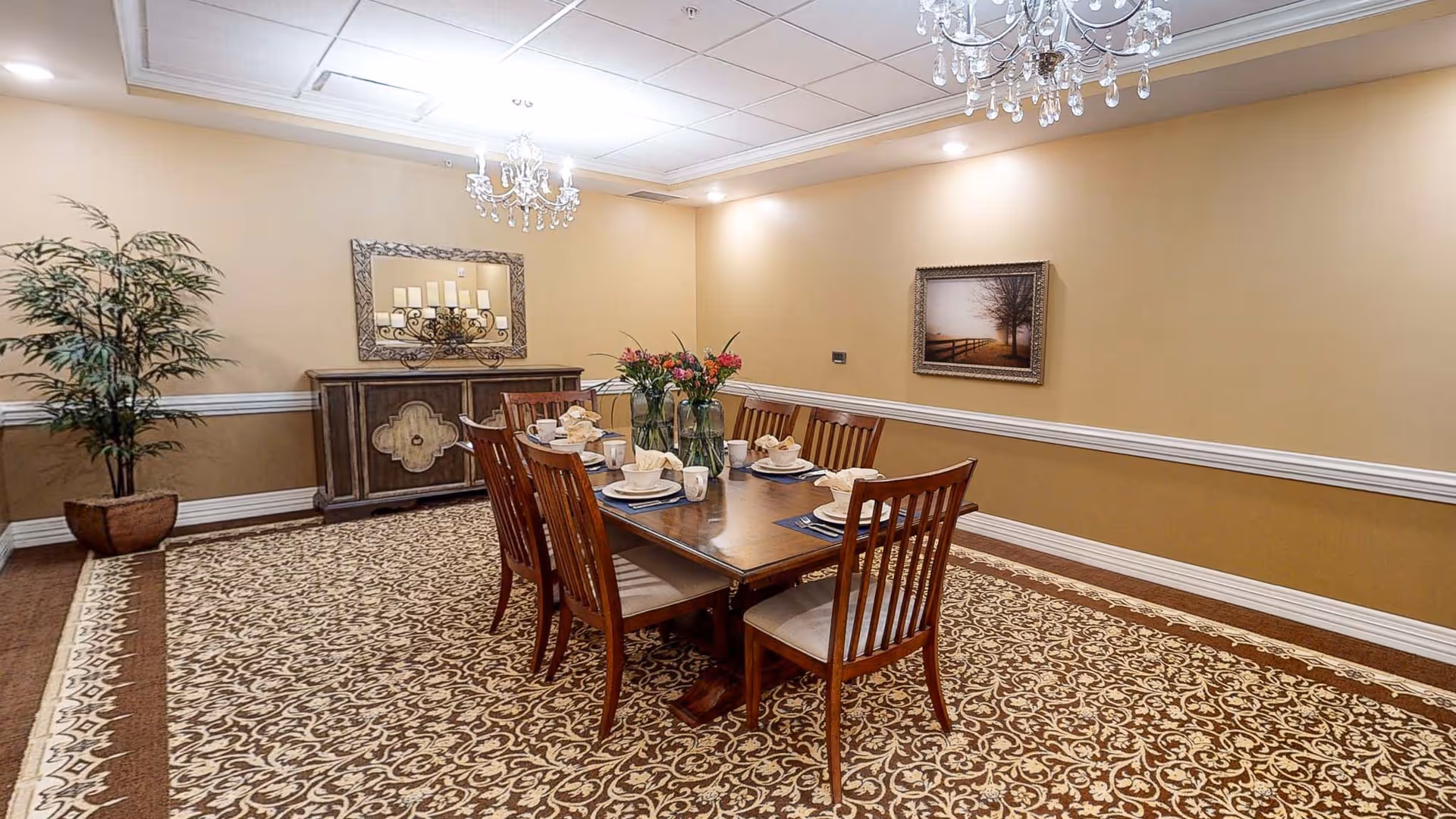 A formal dining room with a long wooden table set for six people with plates, cups, and napkins. The room features a patterned carpet, beige walls with white trim, two chandeliers, a large decorative mirror above a wooden sideboard, a framed picture on the wall, and a potted plant in the corner.