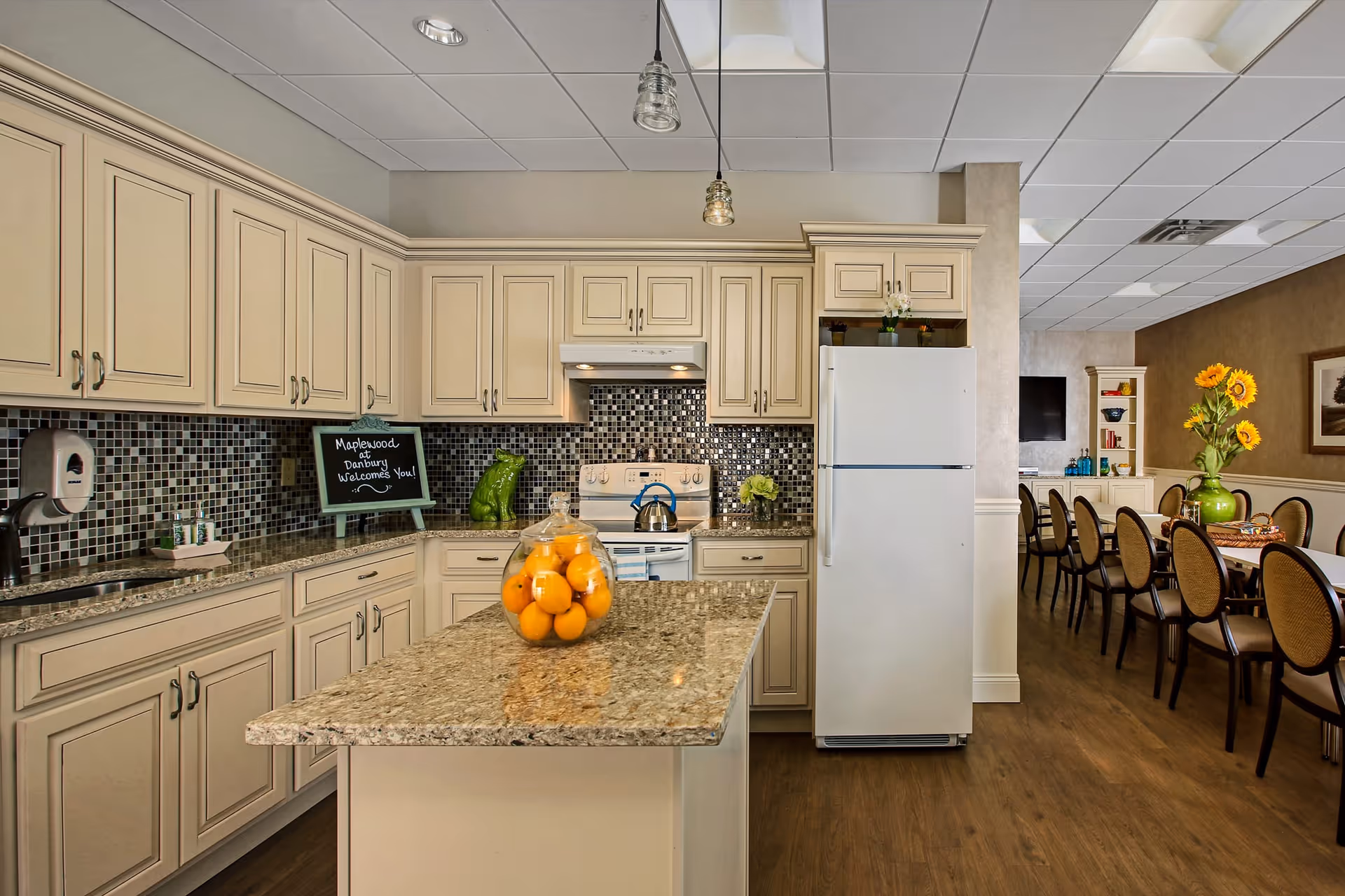 A bright kitchen area with cream-colored cabinets, granite countertops, and a mosaic tile backsplash. A white refrigerator and stove are visible, along with a kitchen island topped with a glass jar filled with oranges. In the background, there is a dining area with a long table, chairs, a vase with sunflowers, and a wall-mounted TV. A small chalkboard on the counter reads 'Maplewood at Danbury Welcomes You!'