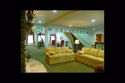 Interior view of a senior living facility lounge area with two yellow sofas arranged around a small table with a lamp. In the background, there is a staircase with a green carpet leading to an upper floor, and several chairs and tables near windows with red curtains. The space is well-lit with ceiling lights and decorated with plants and framed artwork above the staircase.