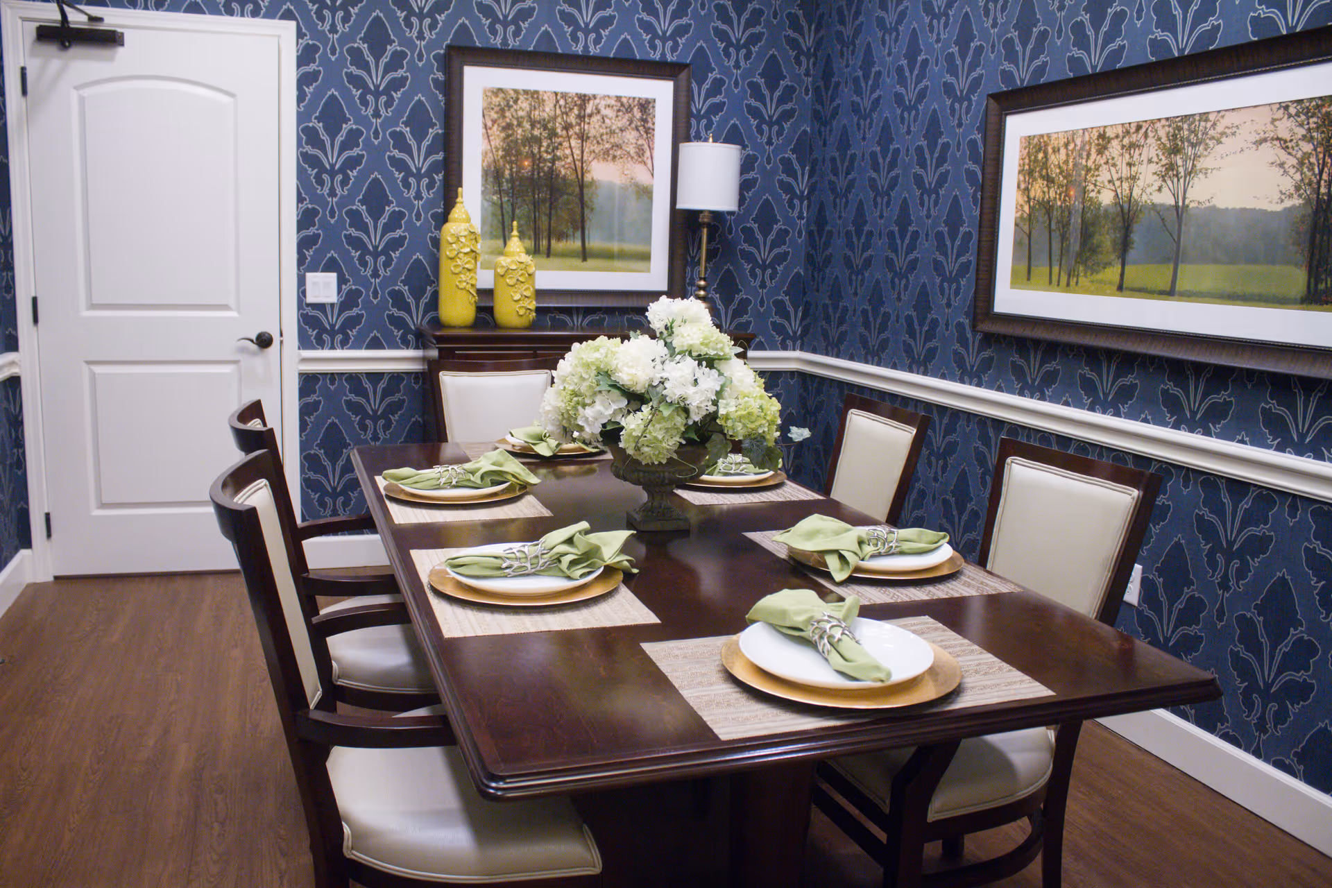 A dining room with a dark wooden table set for six people with white plates, green napkins, and placemats. The room has blue patterned wallpaper, two framed landscape paintings, a white door, a floor lamp, and two yellow decorative vases on a sideboard.