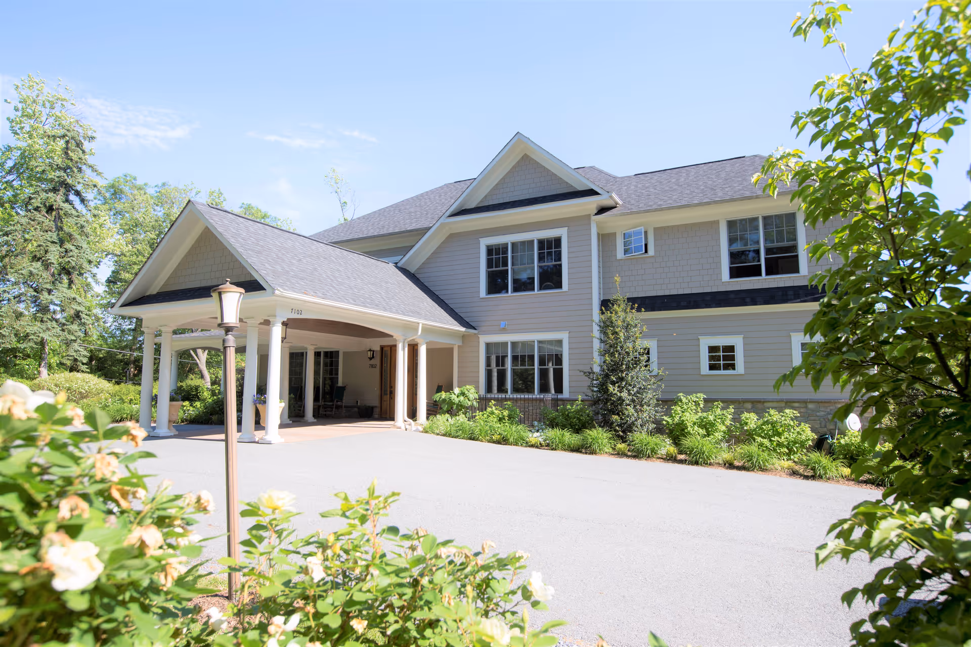 Exterior view of a two-story residential building with a covered entrance supported by white columns, surrounded by greenery and flowering plants under a clear blue sky.