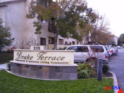 Outdoor view of the entrance sign for Drake Terrace Independent & Assisted Living Community with a parking lot and several parked cars in the background, surrounded by trees and grass.