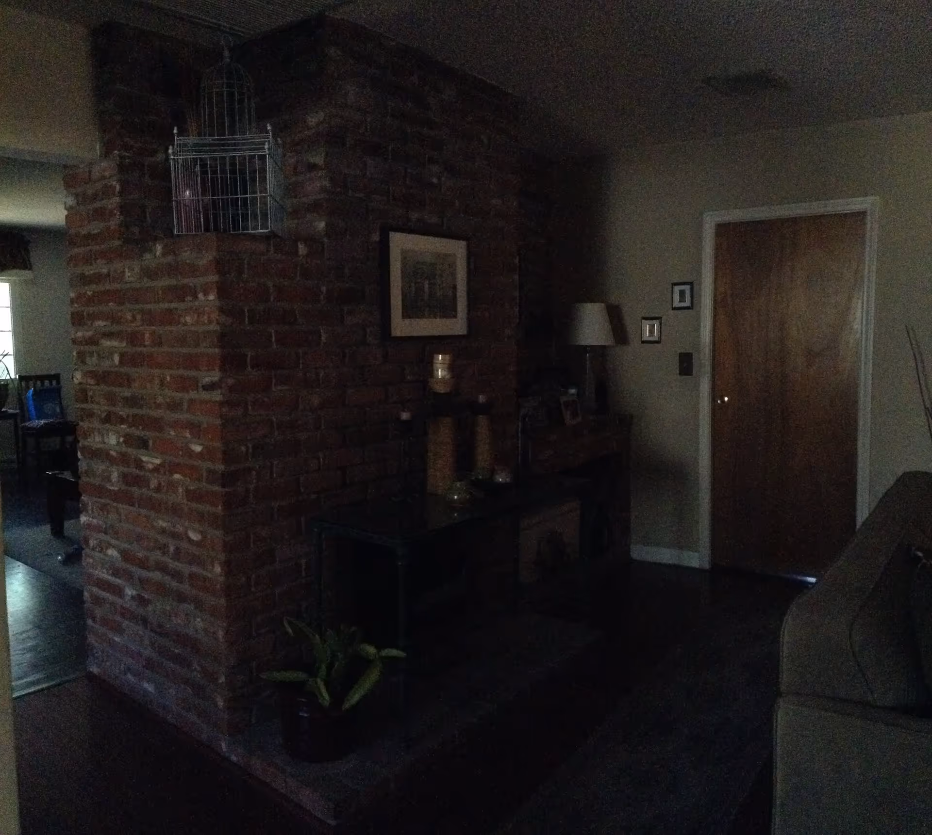 Dimly lit living room featuring a central exposed brick column, a sofa to the right, a console table with decor, and a wooden door at the back.