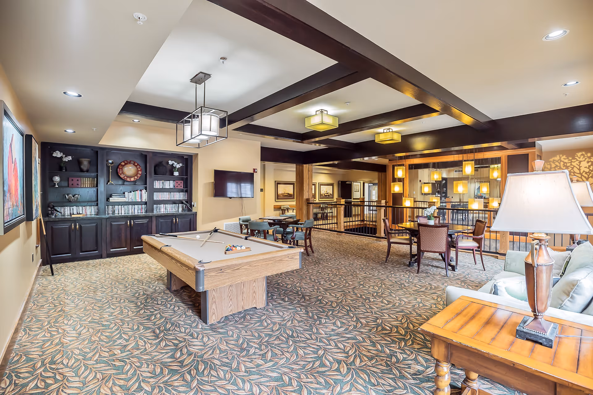 A spacious senior living common area featuring a pool table in the center, a built-in dark wood bookshelf filled with books and decorative items, several tables with chairs for games or socializing, and a cozy seating area with a lamp and couch. The room has patterned carpet, ceiling beams with modern light fixtures, and warm lighting throughout.