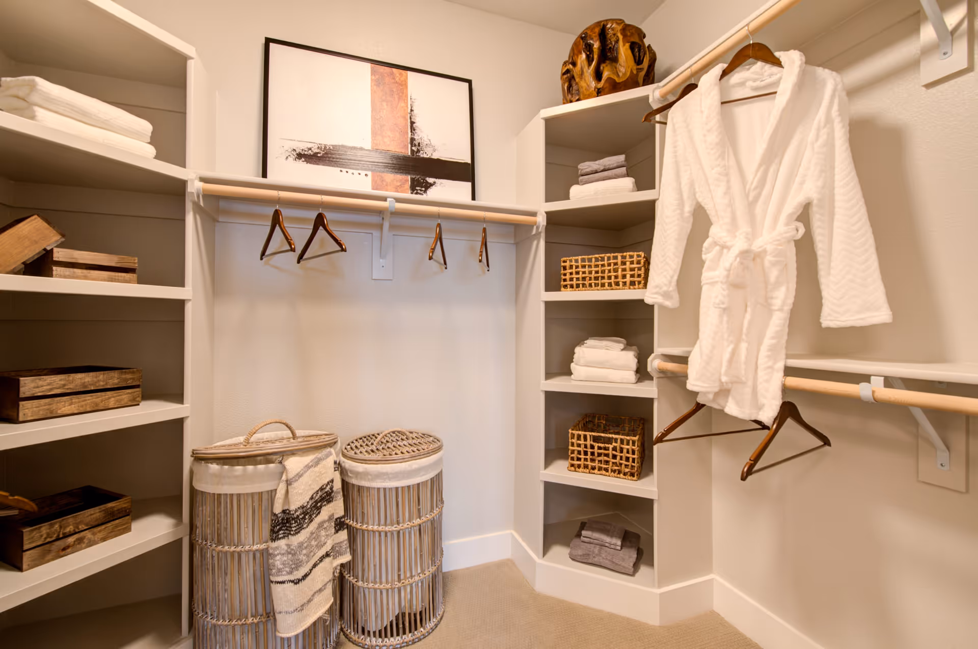 A neatly organized walk-in closet with white shelves holding folded towels, wicker baskets, and wooden boxes. Two wicker laundry hampers with a striped towel draped over one are on the floor. A white bathrobe hangs on a wooden hanger from a wooden rod. A framed abstract artwork is mounted on the wall above the hanging rod.