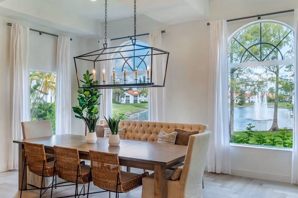 A bright dining room with a large wooden table surrounded by a tufted bench, two upholstered chairs, and three wicker chairs. The room features large arched windows with white curtains, offering a view of a lake with a fountain and greenery outside. A modern rectangular chandelier with candle-style lights hangs above the table, and there are two potted plants on the table.